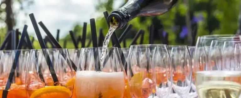 Es wird Sekt in zahlreiche vorbereitete Gläser mit sommerlichen Aperitif-Getränken, Eiswürfeln, Orangenscheiben und schwarzen Strohhalmen bei einer Feier im Freien eingeschenkt.