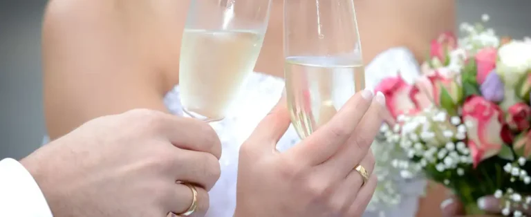 Nahaufnahme der Hände eines Brautpaares, das mit zwei Sektgläsern anstößt. Die goldenen Eheringe sind deutlich sichtbar und die Braut hält im Hintergrund einen Brautstrauß mit rosa und weißen Rosen.