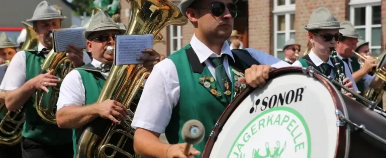 Eine traditionelle Blaskapelle in grünen Westen und grauen Trachtenhüten musiziert mit Instrumenten wie Trommel und Tuba bei einem Festumzug auf einem Schützenfest.