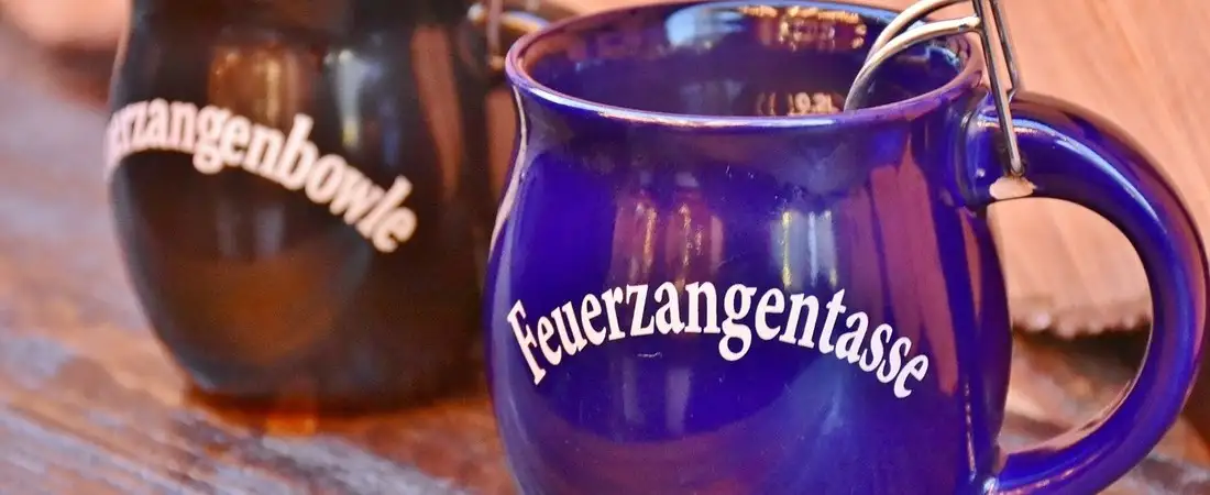 Eine blaue und eine schwarze Keramiktasse mit den Aufschriften "Feuerzangentasse" und "Feuerzangenbowle" stehen auf einem rustikalen Holztisch, passend zu einer winterlichen Veranstaltung.