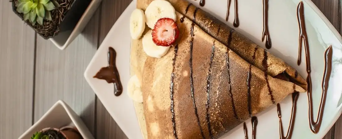 Ein frisch gebackener, gefalteter Crêpe auf einem weißen Teller, ansprechend garniert mit Bananenscheiben, einer Erdbeere und feinen Streifen aus Schokoladensoße.