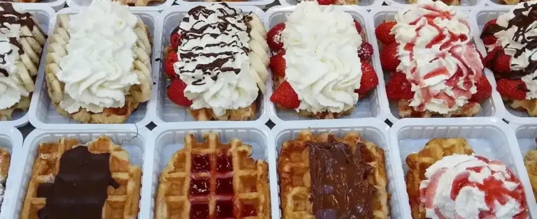 Eine verlockende Auswahl an frisch gebackenen belgischen Waffeln in kleinen weißen Schalen, dekoriert mit vielfältigen Toppings wie frischen Erdbeeren, Bananenscheiben, reichlich Sahne und Schokoladensoße.
